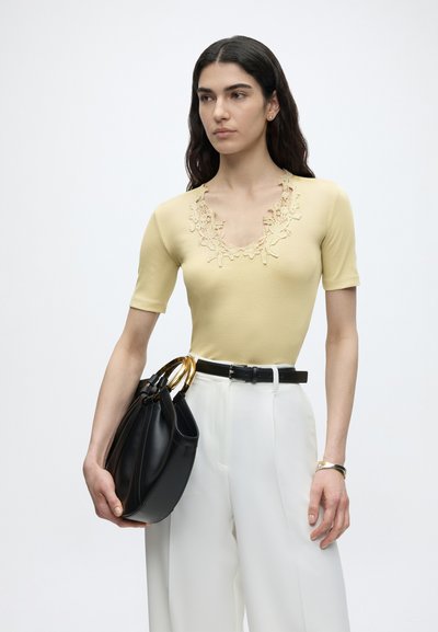 Γυναίκα με μακριά σκουρόχρωμα μαλλιά, που φοράει κίτρινο τοπ με δαντελωτή λαιμόκοψη και λευκό παντελόνι, κρατώντας μια μαύρη τσάντα με χρυσές λαβές.