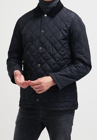 Man som bär en svart vatterad jacka med tryckknappar och fickor med flik, ihop med mörka jeans och en grå polotröja under.