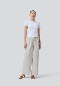 T-shirt bianco a maniche corte abbinato a pantaloni beige chiaro a gamba larga realizzati in un tessuto texturizzato e sandali con tacco marroni. Sfondo semplice.