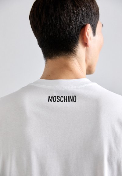 Baltos medvilnės marškinėliai su apvaliu iškirpimu, ant nugaros, žemiau apykaklės, atrodo juodas "MOSCHINO" logotipas.