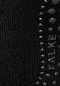 La tela de punto negra presenta un patrón texturizado con puntos grises elevados y "FALKE" impreso en una tipografía elegante a lo largo del costado.