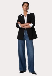 Zwarte dubbelgeknoopte blazer over een wit collared shirt, gecombineerd met losse blauwe high-waisted jeans en zwarte puntige schoenen.