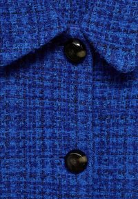 Modra teksturirana tkanina z tkano strukturo in izstopajočim črnim gumbom. Ima ovratnik in gladko teksturo z rahlim sijajem.