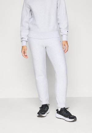 Persona con pantalones deportivos gris claro y sudadera a juego con zapatillas negras y blancas de pie sobre un fondo claro y simple.