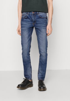 Jeans de ganga azul de corte ajustado com um design clássico de cinco bolsos e costura contrastante. Combinados com sapatos pretos de atacadores.