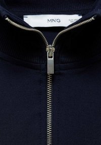 Chaqueta de cremallera azul marino con cremallera metálica, cuello acanalado y textura de tela suave. Incluye una etiqueta de la marca MNG en el escote.