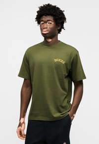 Πράσινο ελαιώδες βαμβακερό t-shirt με κοντά μανίκια, στρογγυλή λαιμόκοψη και κεντημένο λογότυπο "Dickies" σε χρυσό στο αριστερό στήθος. Λεία υφή, άνετη εφαρμογή.