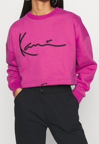Sudadera corta rosa con logo negro bordado, cuello redondo y puños acanalados. Combinada con pantalones negros con bolsillos laterales.