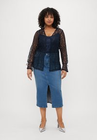 Blusa in pizzo navy con maniche lunghe e fiocco al collo, abbinata a una gonna a tubino di jeans con spacco frontale e orlo rifinito.