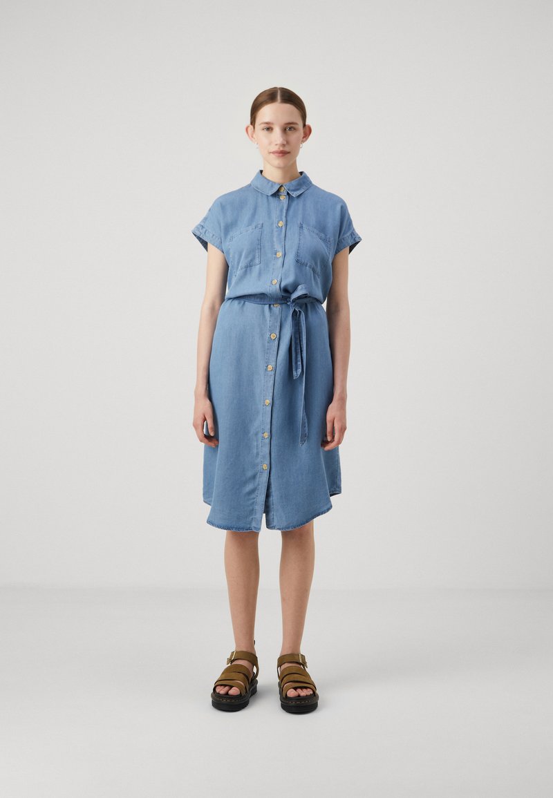 Ljusblå denimskjolklänning med korta ärmar, knäppning fram, två bröstfickor, midjebälte och knälång design. Kombinerad med bruna sandaler.