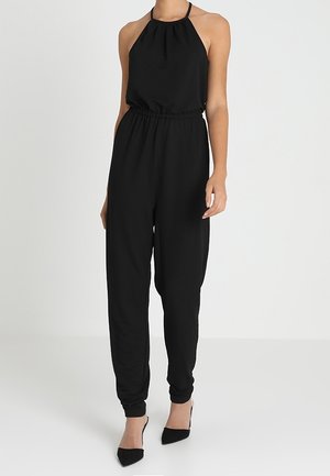 Kobieta ubrana w czarny kombinezon na ramiączkach, z marszczoną talią i zwężanymi nogawkami, w połączeniu z czarnymi szpilkami o spiczastym nosku.