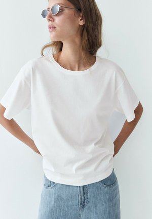 Femme portant des lunettes de soleil rondes, un t-shirt blanc à manches courtes et un jean bleu clair, debout les mains sur les hanches devant un fond blanc.