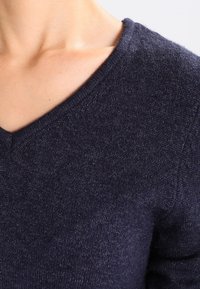 Mörkblå, stickad tröja med en mjuk textur och V-ringning. Höjdpunkter inkluderar subtila fläckar och en åtsittande form.