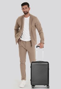 Valigia rigida nera con design ondulato testurizzato, manico retrattile e quattro ruote. Affiancata a un uomo in abbigliamento casual beige.