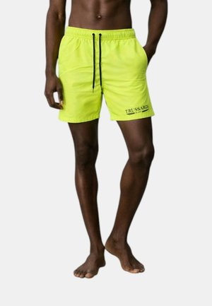 Uomo con pantaloncini da bagno giallo brillante con lacci neri e logo "Trussardi" sulla gamba sinistra, in piedi a piedi nudi su sfondo semplice.