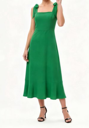 Femme portant une robe midi verte sans manches avec des bretelles nouées et un ourlet volanté, associée à des sandales noires à talons hauts avec bride à la cheville.