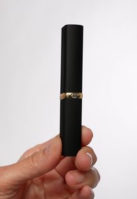 Svart cylindrisk läppstiftkapsel med matt yta och ett guldband med präglade detaljer runt mitten. Hand som håller produkten.