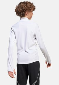 T-shirt de sport à manches longues, blanc, avec une coupe ajustée et un col montant, côtés texturés, associé à un pantalon noir avec des accents blancs.