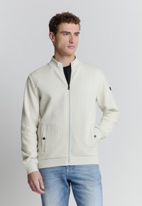 Lichtcrème zip-up jack met een gewatteerde textuur aan de voorkant, effen mouwen, twee zijzakken met zwarte accenten en een staande kraag.