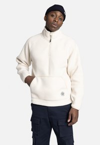 Crème fleece pullover met een hoge kraag en een kwart rits. Heeft een voorzak, ribgebreide manchetten en een rond logo aan de onderkant.