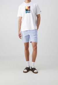 T-shirt blanche avec un imprimé graphique, associée à un short rayé bleu clair et blanc, des chaussettes blanches et des sandales foncées.
