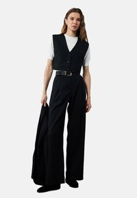 Zwarte op maat gemaakte vest met een V-hals, knoopsluiting, gedragen over een wit t-shirt met korte mouwen. Zwarte broek met wijde pijpen en een bijpassende blazer.