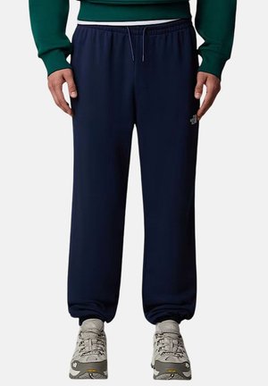 Uomo che indossa pantaloni jogger blu navy con vita elasticizzata, maglia verde a maniche lunghe e scarpe da trekking bianche e grigie, in piedi contro uno sfondo bianco.
