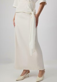 Longuette beige con una texture liscia, dotata di una cintura bianca legata e borchie dorate lungo la vita. Abbinata a décolleté crema con punta aogni.