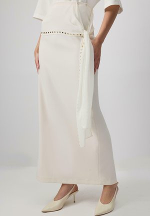 Longuette beige con una texture liscia, dotata di una cintura bianca legata e borchie dorate lungo la vita. Abbinata a décolleté crema con punta aogni.