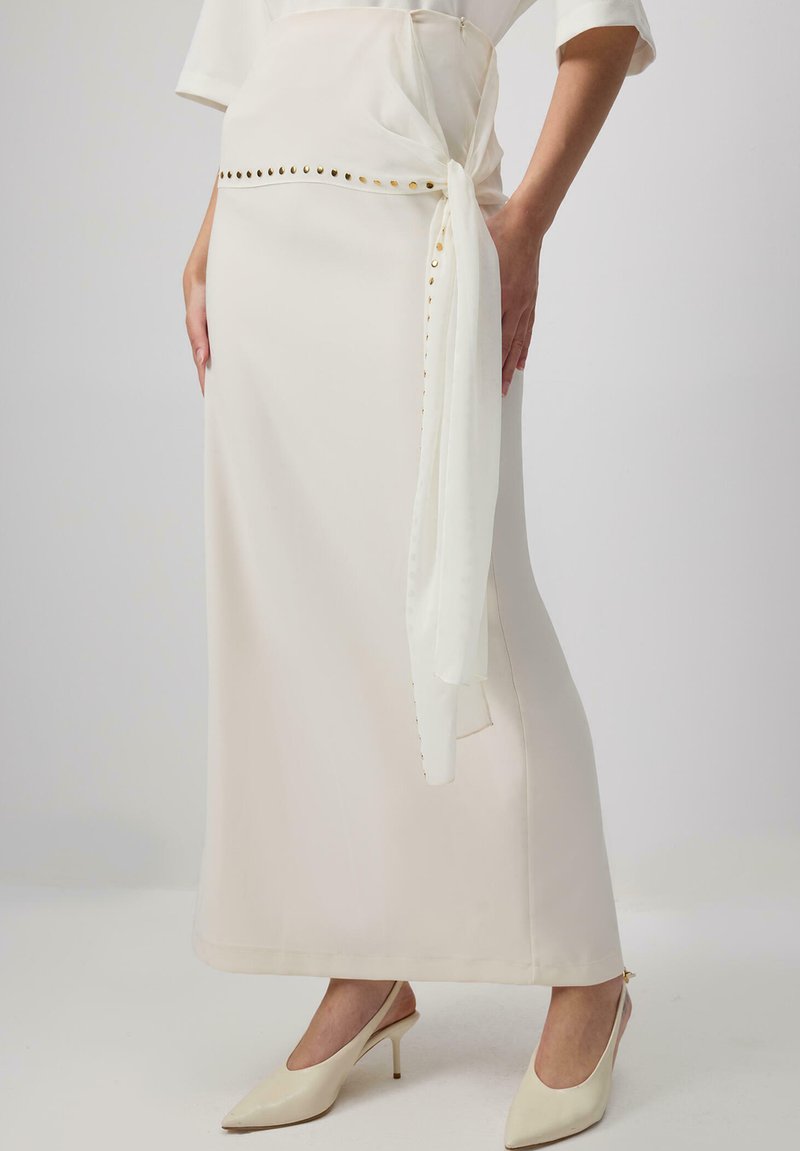 Longuette beige con una texture liscia, dotata di una cintura bianca legata e borchie dorate lungo la vita. Abbinata a décolleté crema con punta aogni.