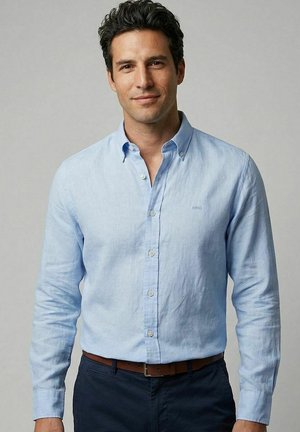 Man with dark hair wearing light blue button-up shirt and dark pants standing against a plain light gray background.