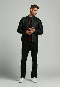 Man in black leather jacket, dark pants, white sneakers standing against plain gray background, looking down and adjusting jacket.