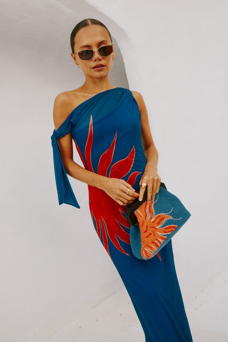 Abito blu a spalla unica con un design a raggi di sole rossi; abbinato a una borsa a tracolla testurizzata blu e arancione. Includono occhiali da sole e gioielli.