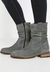 Des bottines en suède gris présentent un design décontracté avec une fermeture éclair sur le côté, un talon en bois marron et de subtils accents de coutures sur les côtés.