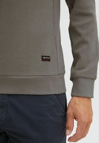 Szary, teksturowany sweter z dopasowanym krojem i prążkowanymi mankietami, z małą metką marki po dolnej stronie. Uzupełniony o granatowe spodnie.