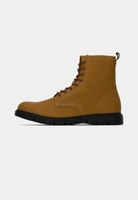 Beżowe zamszowe botki za kostkę mają brązowe sznurowadła, pętelkę do wciągania oraz wytrzymałą czarną gumową podeszwę z widocznym bieżnikiem.