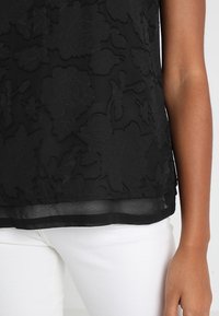 Svart blommig broderad topp med en genomskinlig textur, med en solid svart fåll. Kombinerad med vita jeans som visar en lagerdesign.