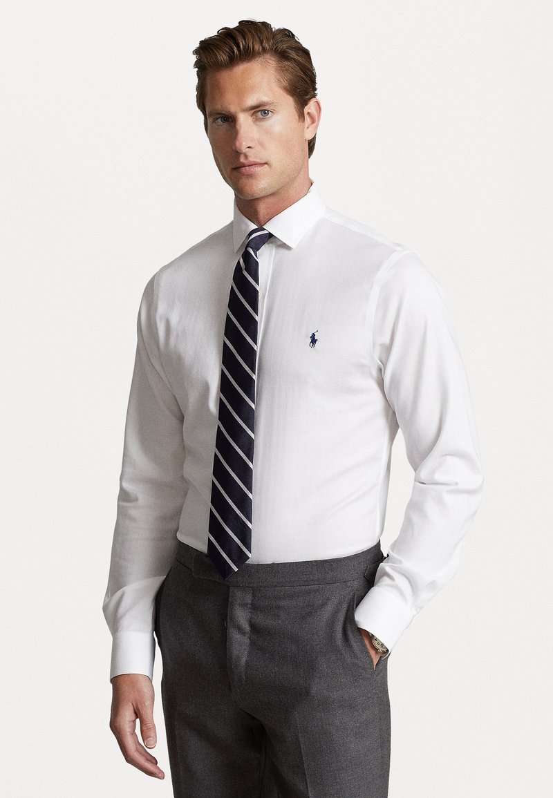 White long-sleeve shirt with a small blue logo on the left chest, paired with a navy and white striped tie and dark gray trousers.