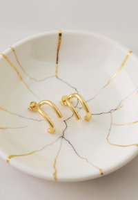 Orecchini in oro con un design curvilineo appoggiati su un piatto bianco con motivi di marmo neri e dorati. Finitura elegante e superficie liscia.