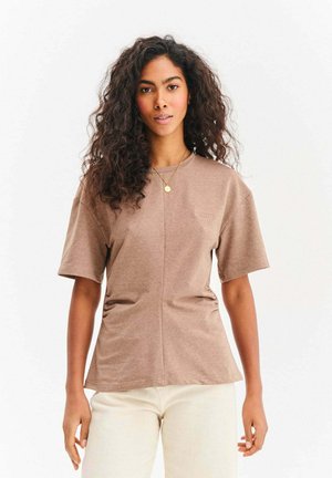 Kobieta o długich kręconych włosach, ubrana w brązową koszulę z krótkim rękawem i białe spodnie, stojąca na tle jednolitego białego tła.