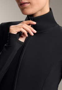 Black high-collared garment with a zipper, made of smooth, stretchable material. Close-up of hand adjusting the zipper near the neck.