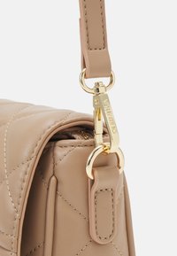 Vadderad beige handväska i konstläder med guldfärgade metallbeslag och clips. Har en platt rem och detaljerad sömnad längs kanterna.