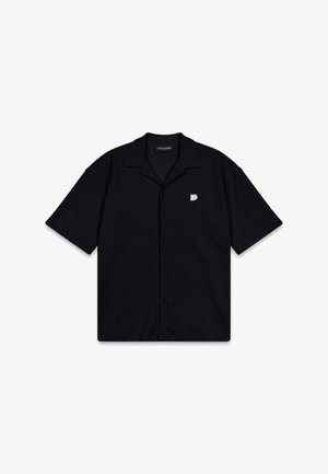 Camisa negra de manga corta con botones y cuello, con un pequeño logo blanco en forma de "P" en el pecho izquierdo, extendida sobre un fondo blanco.