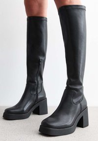 Botas de cuero negro hasta la rodilla con un tacón grueso, cremallera lateral, textura suave y detalles de costura mínimos.