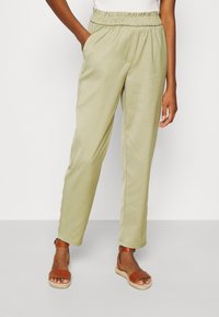 Lichtgroene elastische broek met zijzakken, taps toelopende pijpen en een gladde, lichtgewicht stof. Gepaarde met bruine sandalen met banden.