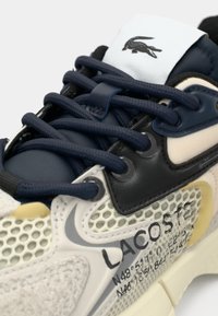 Sneaker Lacoste con una toma in rete crema, dettagli in blu navy e particolari neri testurizzati. Presenta lacci rotondi e una patch con il logo sulla linguetta.