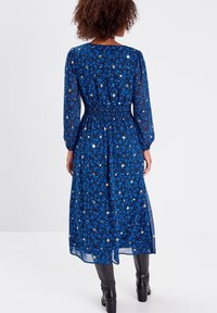 Robe longue bleue avec motif floral, manches longues transparentes, taille cintrée et jupe fluide. Associée à des bottes noires montantes.