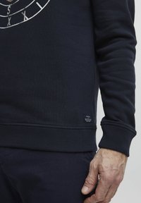 Námořnická modrá mikina s žebrovanými manžetami a lemem, kulatým výstřihem a malou nálepkou na dolním okraji, hladká textura.