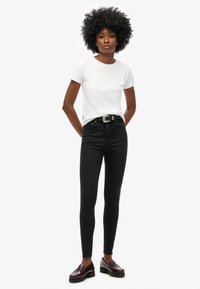 Witte korte mouwen t-shirt gecombineerd met zwarte high-waisted skinny jeans, geaccentueerd door een zilveren riem en donkere loafers.
