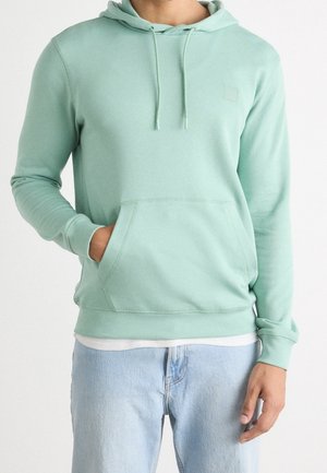 Person trägt einen hellgrünen Kapuzenpullover mit Fronttasche und hellblaue Jeans, steht mit einer Hand in der Tasche.